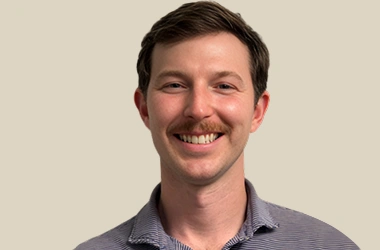 Professional headshot of smiling male dentist in purple shirt against neutral background.