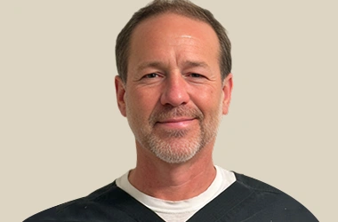 Professional headshot of a man smiling at camera in casual attire against neutral background.